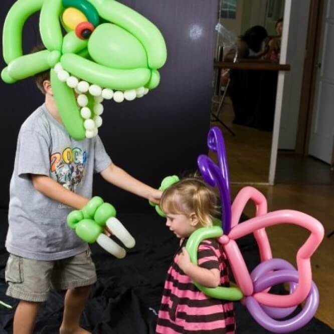 Child wearing butterfly balloon costume interacts with another child in a large green dinosaur balloon outfit.