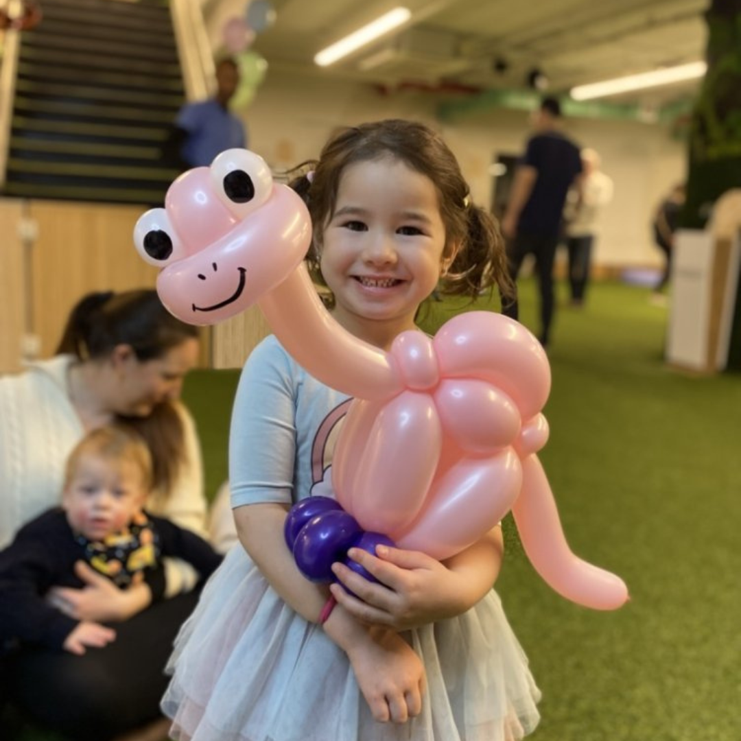 Smiling young girl holding a pink balloon dinosaur sculpture at a fun balloon party with Dave Brenn.
